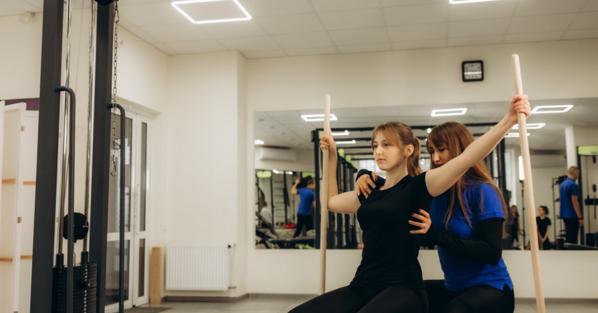 A woman going through scoliosis physical therapy. She is holding two wooden rods while another woman supports her back.
