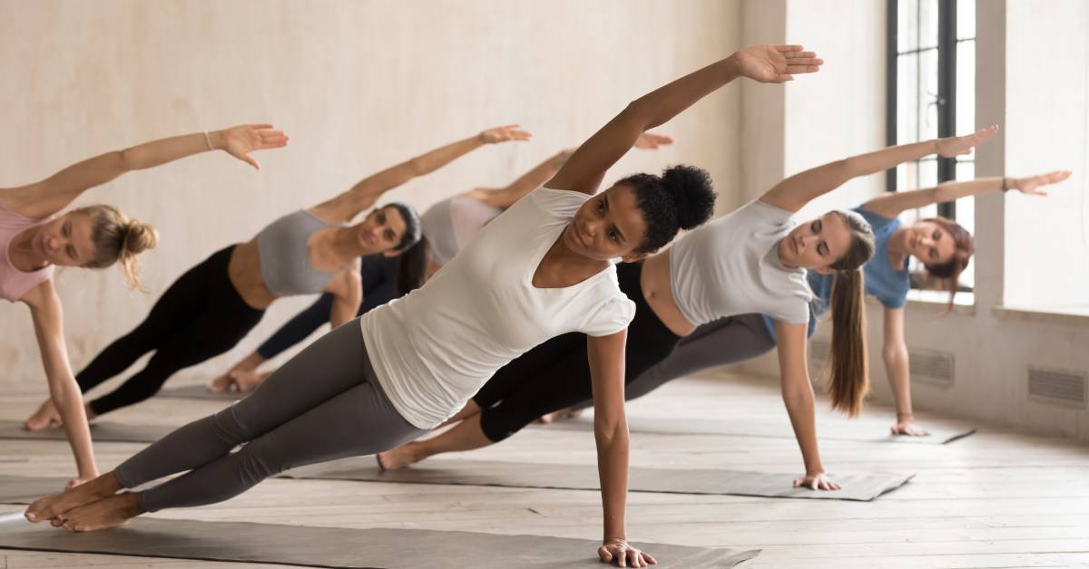 A group of people performing side plank poses on mats in a bright studio with natural light coming through windows.