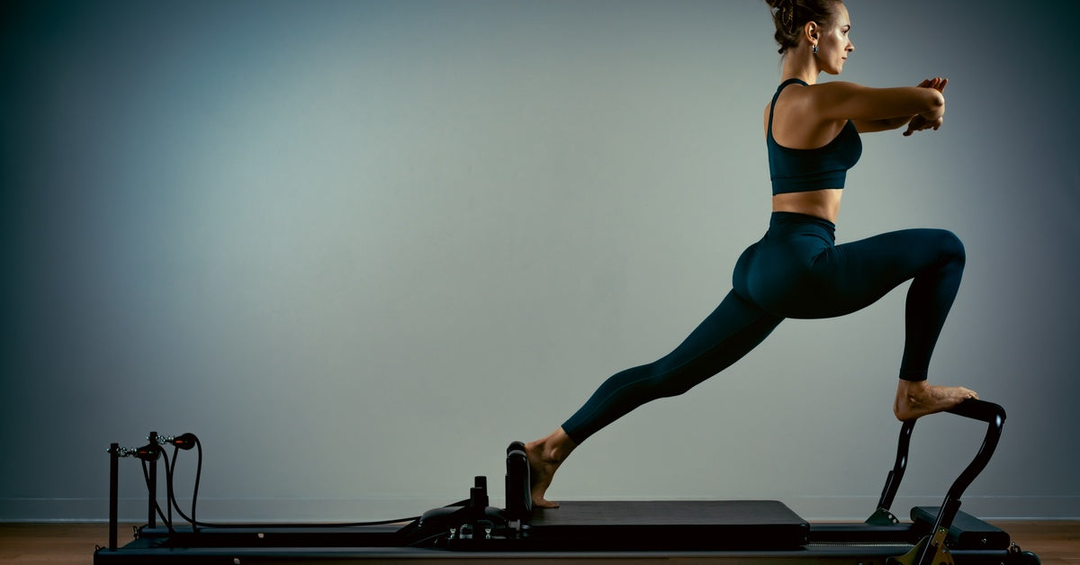 A Young woman with her arms up and legs in a lunge is exercising on a Pilates reformer bench in front of a grey wall.