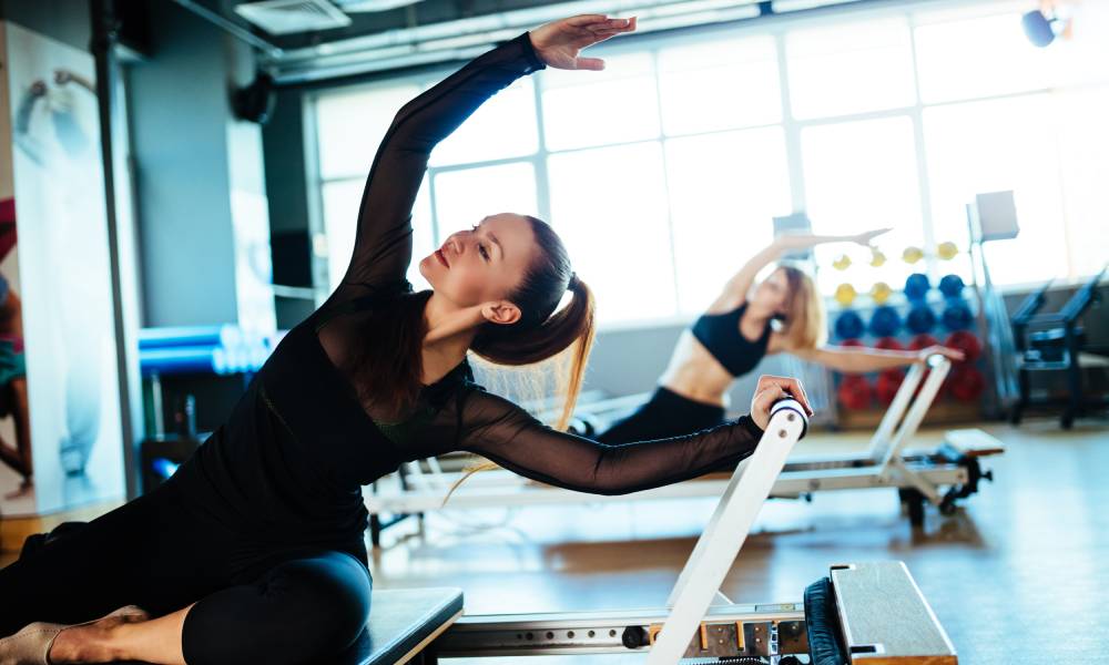 Two young women completing Pilates exercises on pieces of gym equipment. They appear focused and mindful of their exercises.