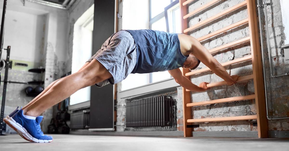 A man wearing a tank top and shorts performs an exercise against a mounted Pilates wall bar during a workout.