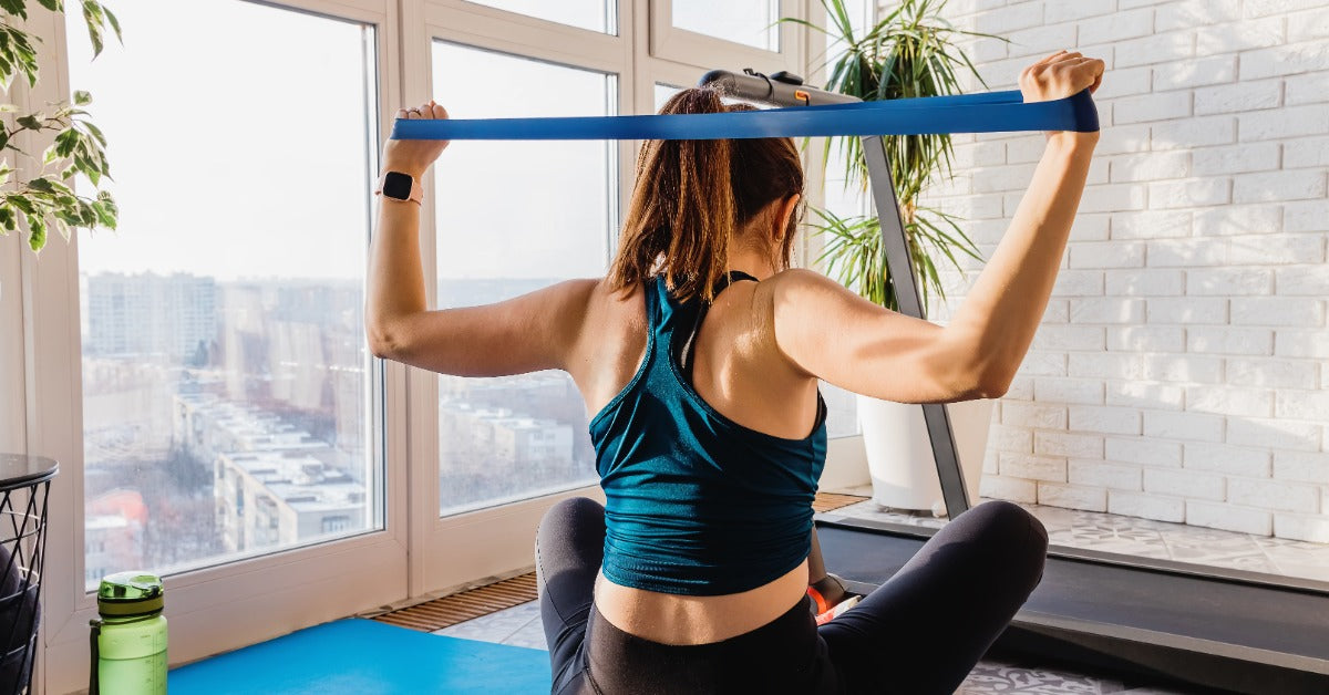 A girl stretching a resistance band while sitting on an exercise mat in a bright room with wood flooring.